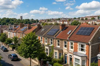A realistic wide-angle image of a Bristol residential street with terraced and semi-detached houses, showing discreet roof-mounted solar PV panels on clay-tiled roofs under bright but partly cloudy UK daylight. Include typical local details such as chimneys, rooflights, mature trees and varied roof orientations, with no logos, people, branding or text overlays.