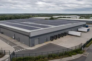 A realistic wide photograph of a large UK logistics warehouse on an industrial estate under soft overcast daylight, with rows of solar panels across a broad flat roof, low-pitch…
